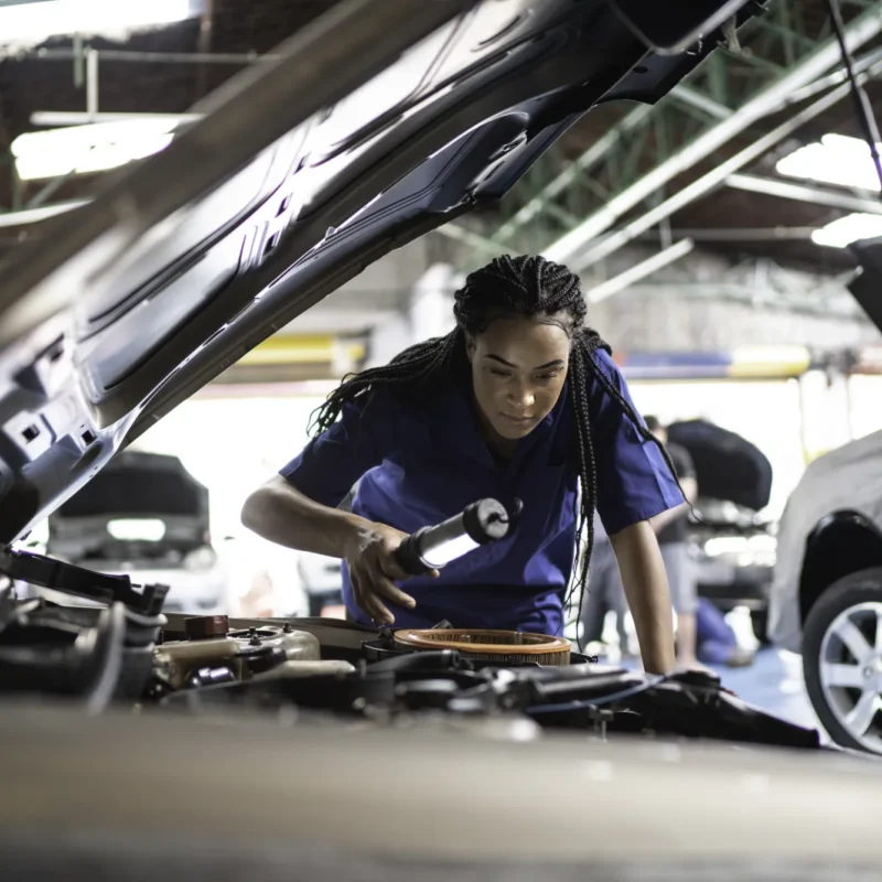 A mechanic in a blue uniform shines a flashlight into a car's engine bay under the raised hood inside an auto repair shop.