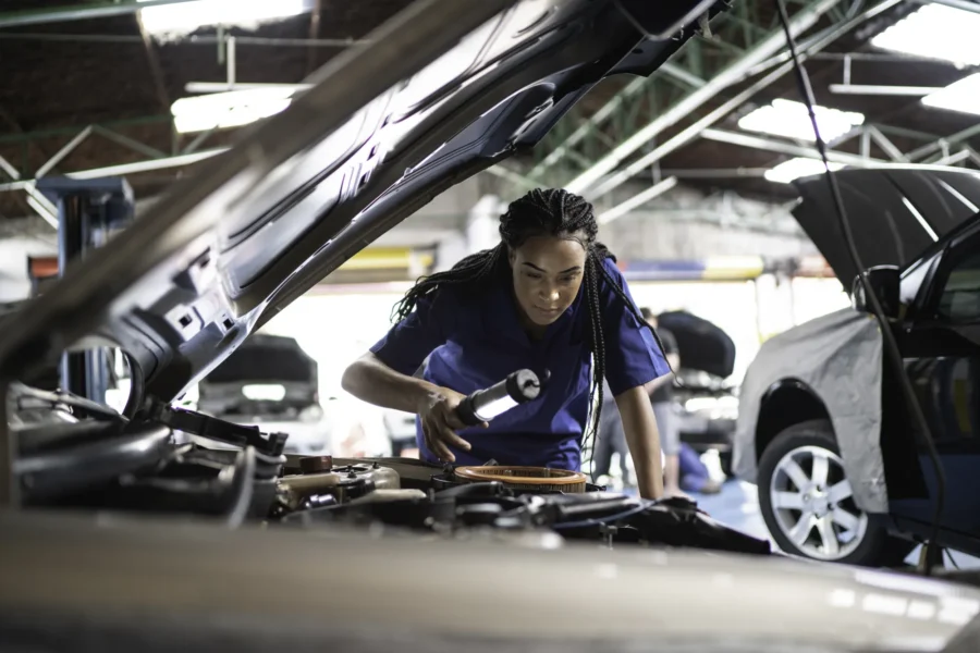 A mechanic in a blue uniform shines a flashlight into a car's engine bay under the raised hood inside an auto repair shop.