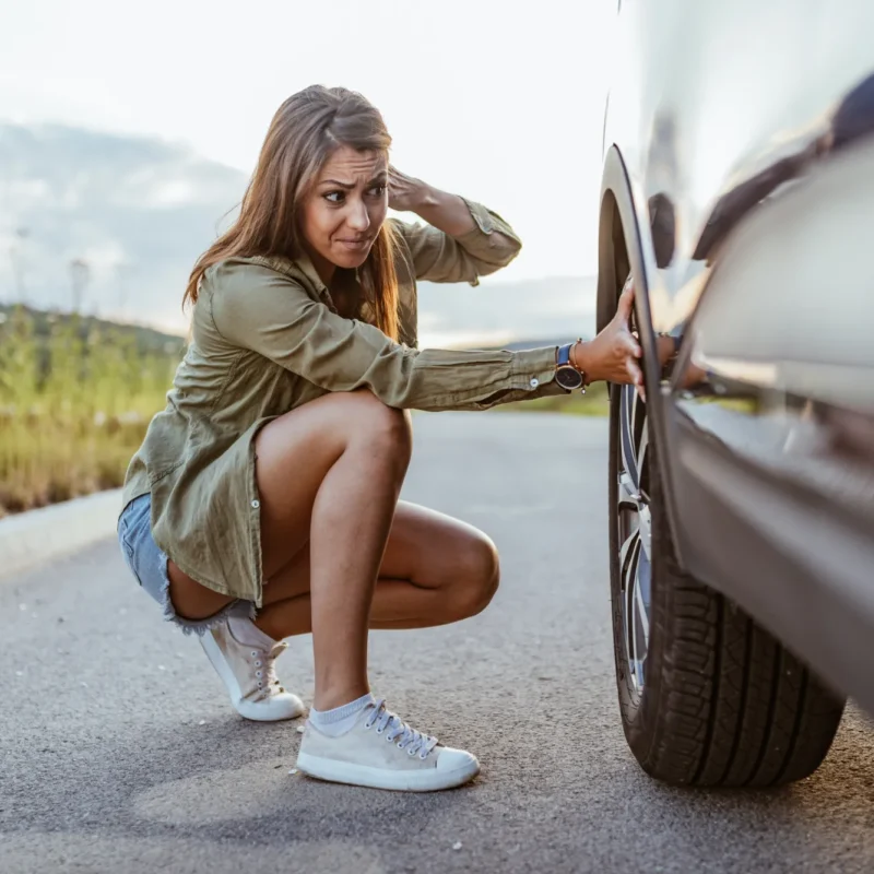 A worried woman crouches next to the front tire of her car on the side of a road.