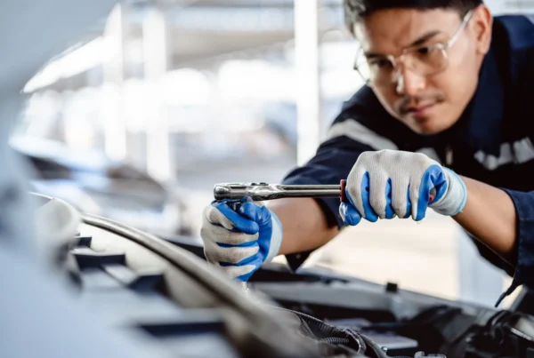 A mechanic wearing safety glasses and work gloves uses a ratchet wrench on an engine under an open car hood.