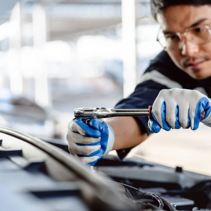 A mechanic wearing safety glasses and work gloves uses a ratchet wrench on an engine under an open car hood.
